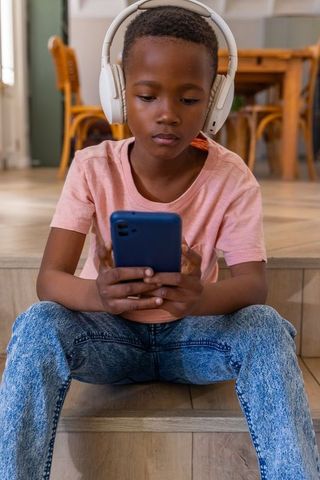 African american boy sitting on stairs listening with headphones while using smartphone