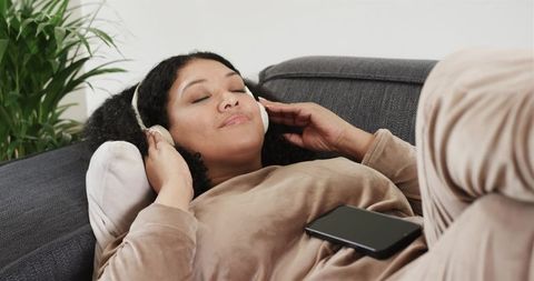 Woman relaxing on gray sofa wearing headphones with smartphone on torso