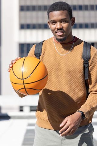 Smiling african american man holding basketball on rooftop with backpack and smartwatch