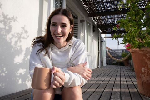Smiling woman enjoying coffee on wooden veranda under sunlit pergola