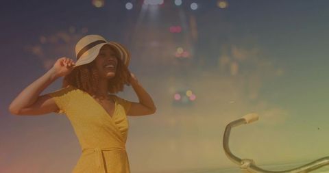 Smiling woman in yellow dress adjusting straw hat by seaside with bike handlebar at golden hour