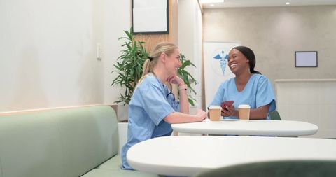 Two nurses laughing and sharing coffee while sitting in modern clinic breakroom with stethoscope