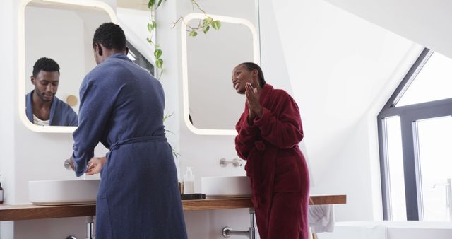 African American couple enjoying their morning routine in a modern bathroom. The man is washing his hands at a sink while the woman stands beside him joyfully in a red bathrobe. Ideal for content related to daily rituals, home life, relationships, personal care, hygiene, and modern household settings.