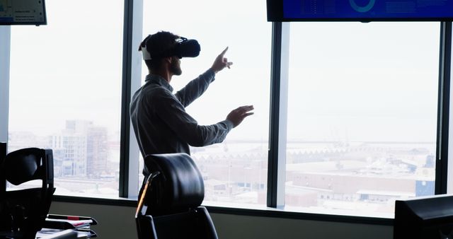 Businessman wearing VR headset in a modern office with large windows. Cityscape visible through windows. Useful for themes related to business innovation, technology in the workplace, future of work, interactive virtual tools, and professional environments.