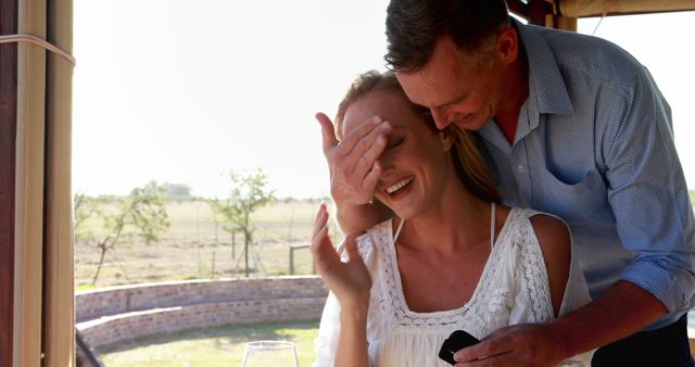 A middle-aged Caucasian man is surprising a woman, his partner, with a ring, suggesting a marriage proposal, with copy space. Their joyful expressions and the intimate gesture create a moment of romance and commitment.