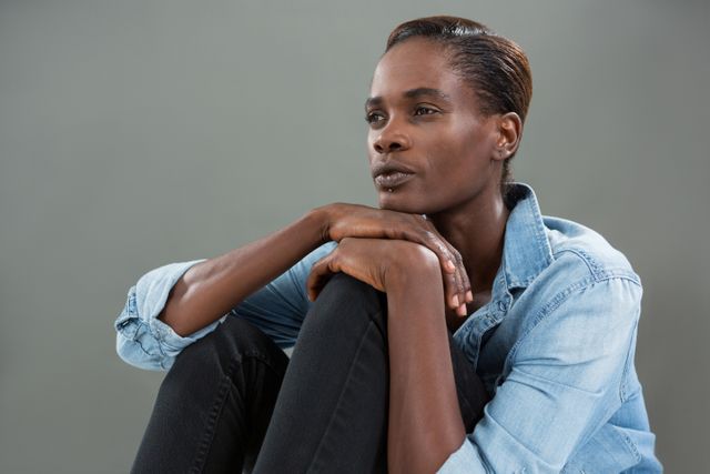 This image depicts an androgynous man in a denim shirt, sitting in a thoughtful pose against a grey background. Ideal for use in articles or advertisements focusing on fashion, modern lifestyle, introspection, or gender fluidity. Perfect for illustrating themes of contemplation, serenity, and casual style.