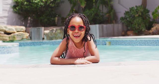 This image showcases a joyful child with braids and red sunglasses smiling next to a pool. It is perfect for use in summer vacation promotions, children's swimwear ads, family travel brochures, and articles about outdoor activities and fun. It conveys a sense of joy, relaxation, and lively energy.