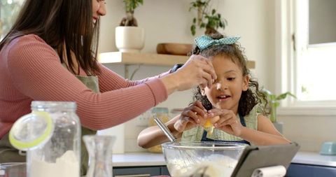 Mother guiding daughter cracking egg while baking together in bright home kitchen