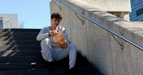 Young man sitting on concrete steps opening paper bag in gray tracksuit with earbuds, sunlit urban m