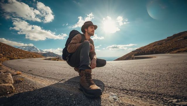 Crouching hiker adjusting hiking boots on winding alpine road, backpack and hat