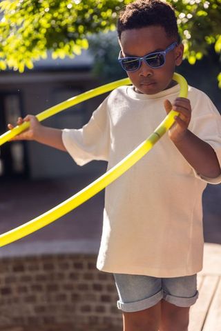African american boy playing with neon yellow hula hoop outdoors wearing blue sunglasses
