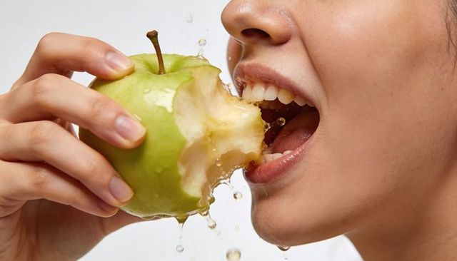 Young woman biting juicy green apple, holding fruit with splashing juice closeup