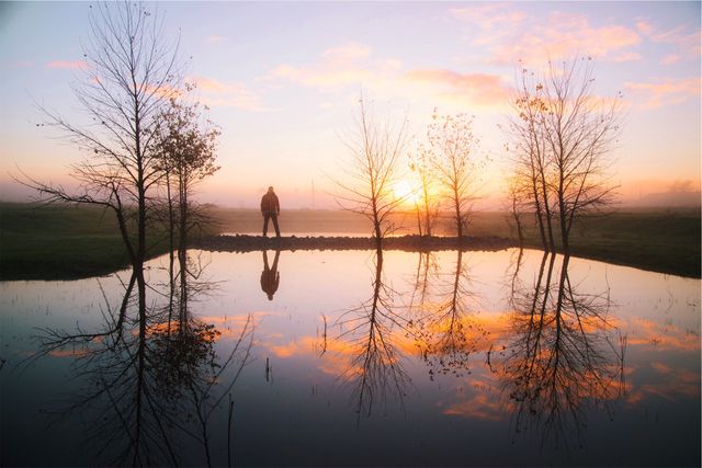 Silhouette of a person standing near a tranquil pond with the beautiful reflection of trees and sky. Ideal for concepts related to peace, solitude, nature, and early morning. Can be used in projects promoting relaxation, meditation retreats, inspirational quotes, and nature conservation campaigns.