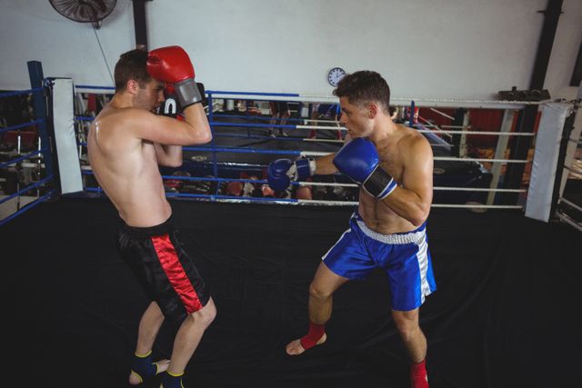 Two boxers are sparring in a boxing ring at a gym. Both athletes are wearing boxing gloves and shorts, engaged in an intense training session. This image can be used for promoting fitness centers, boxing classes, sports events, or articles related to martial arts and physical fitness.