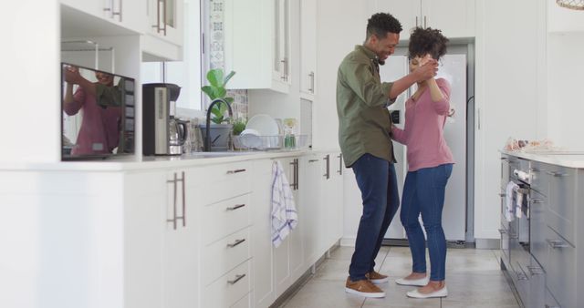 An African American couple enjoys a dance together in a modern, white kitchen, fostering a warm and joyful atmosphere. This image conveys themes of love, togetherness, and happiness in everyday life. It is perfect for advertisements, articles on relationships and lifestyle, and content promoting family or household products.