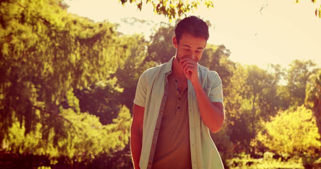 Young man in casual clothing enjoying a moment of reflection in a tranquil park filled with lush greenery. Sunlight filters through the trees, creating a warm and serene atmosphere. Ideal for use in themes related to relaxation, nature, mindfulness, outdoor activities, and tranquility.