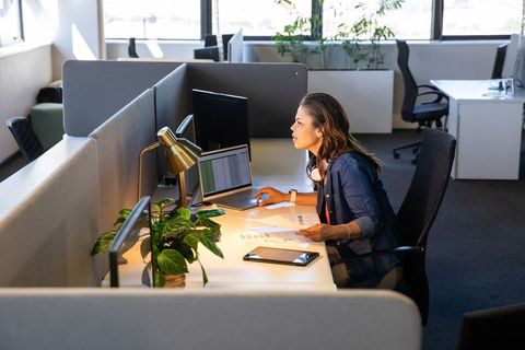 Woman working in modern office cubicle analyzing charts on laptop with headphones