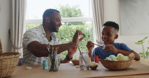 African american father and son painting easter eggs together at sunny kitchen table