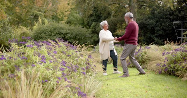 Happy diverse senior couple holding hands and dancing in garden, copy space. Retirement, togetherness, love, romance, fun, nature and active senior lifestyle, unaltered.
