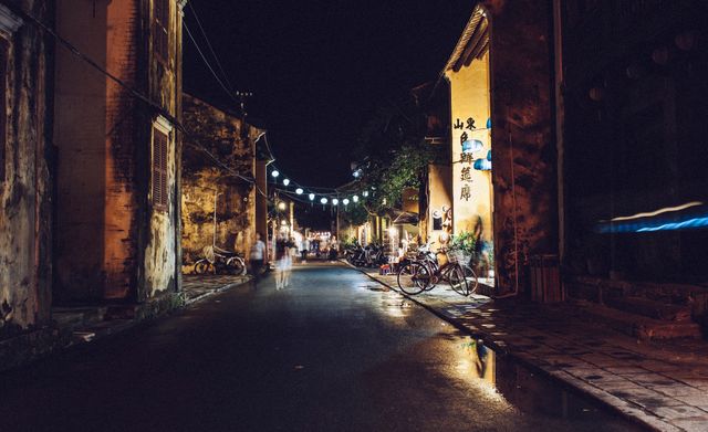 A serene night scene featuring a quiet street illuminated by lantern lights in a charming Asian town. The street has wet pavement reflecting the lights, with bicycles lined along the sides and traditional buildings creating a nostalgic feeling. Ideal for conveying travel inspiration, tranquility, cultural heritage, and nighttime photography.