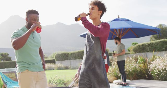 Group of friends enjoying a barbecue party in sunny outdoor setting. The scene depicts people drinking and socializing under a blue umbrella, with a mountainous landscape in the background. Ideal for websites or ads promoting outdoor gatherings, summer activities, or social events.