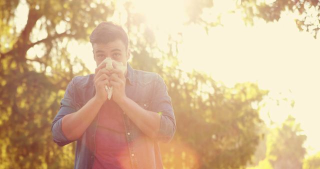 A young Caucasian man is blowing his nose into a tissue outdoors, with copy space. Sunlight filters through the trees behind him, suggesting a seasonal setting that might be related to allergies or a cold.