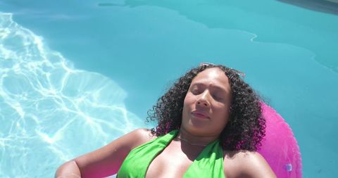 African american woman floating on pink inflatable in pool wearing green swimsuit