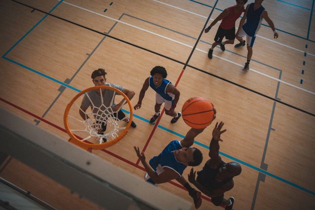 This dynamic image captures an intense moment in a basketball game from an overhead perspective. It shows diverse male players actively engaged in shooting and defending at an indoor court. Ideal for use in sports-related articles, team-building promotions, athletic training materials, and advertisements for sports equipment or facilities.
