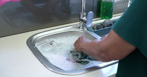 Senior african american woman handwashing laundry at kitchen sink with submerged glasses and suds