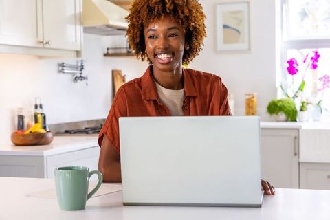 Cheerful african american woman working remotely at sunlit kitchen island with laptop