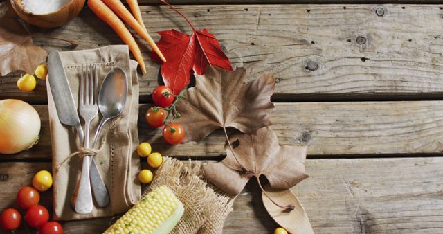 Rustic fall-themed arrangement featuring silver utensils wrapped in burlap, surrounded by fresh vegetables like tomatoes, carrots, and corn on a wooden table. Great for illustrating autumn harvest themes, seasonal dining, promoting healthy eating during fall, and decorating ideas for Thanksgiving.