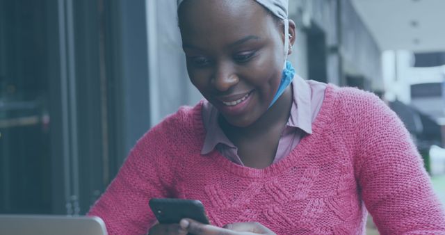 African American woman sitting outdoors, smiling while using smartphone. She is wearing a pink sweater and headwrap, creating a casual look. Suitable for themes related to technology, communication, social media, urban lifestyle, and modern connectivity.