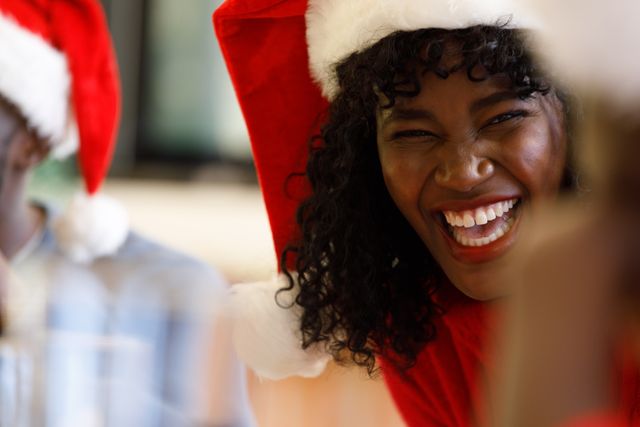 Close up of an african-american woman wearing a santa hat laughing with family at a dinner table. beside her in the background is a man wearing a santa hat