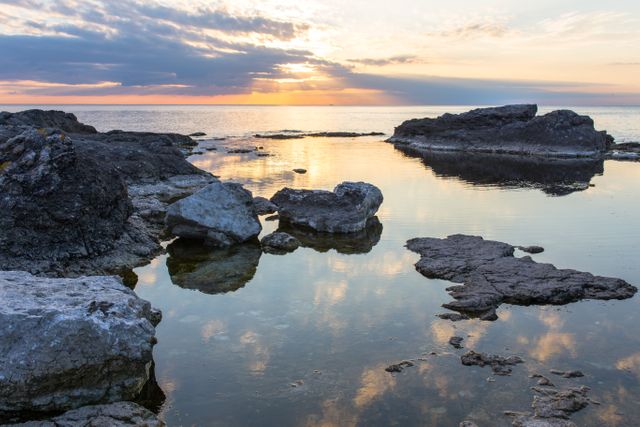 The image shows a tranquil coastal landscape at sunset, with rocky formations reflecting in the calm waters. The sky is filled with clouds, and the sun setting on the horizon adds a warm, golden hue. This stock photo can be used for travel brochures, nature blogs, wallpapers, relaxation apps, and environmental awareness campaigns.