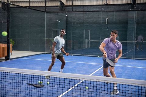 Diverse male partners playing padel on blue court at indoor club, dynamic doubles rally