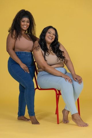 Smiling friends posing on red chair wearing beige tops and jeans on yellow backdrop celebrating conf