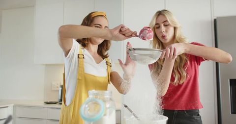 Two friends sifting flour and laughing while baking together in bright kitchen at home