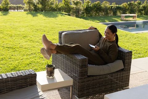 Sunlit asian woman relaxing poolside on wicker chair using smartphone in olive joggers