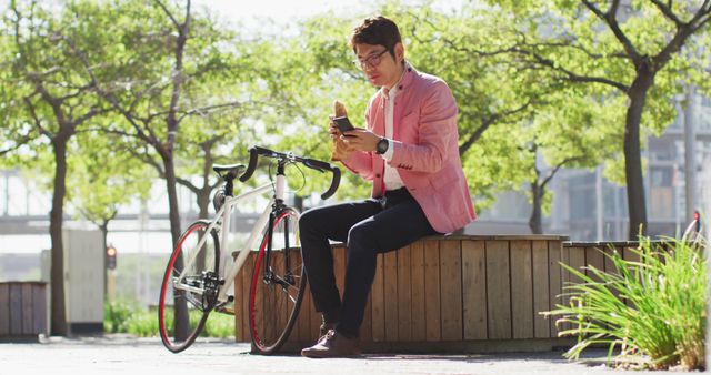 Young man sits on bench in a park, enjoying a croissant and using his smartphone. Dressed in casual clothing, sitting next to a bicycle on a sunny day. Can be used for lifestyle, leisure activities, technology usage, outdoor scenes, or urban living themes.