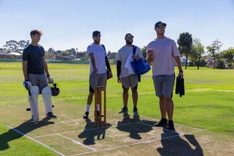 Multiethnic cricket teammates standing on sunlit pitch with stumps and protective pads