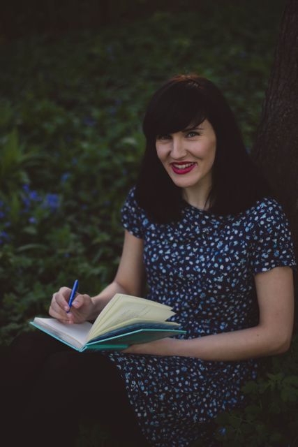 This stock photo depicts a woman sitting under a tree in a park, writing in a notebook. She is smiling, suggesting a sense of relaxation and contentment. The setting appears serene with greenery and flowers around, offering a perfect backdrop for creative inspiration. Ideal for concepts related to journaling, creativity, leisure activities, nature connection, and peaceful moments. Perfect for use in lifestyle blogs, articles related to personal development, and marketing materials for leisure activities.