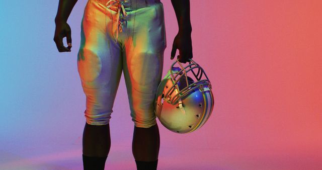 Football player holding helmet under colorful studio lighting, creating a striking visual effect. This image is ideal for sports-related advertisements, fitness campaigns, team spirit promotions, and athletic wear branding. Suitable for websites, social media posts, and magazine covers emphasizing fitness, sports, and athleticism.