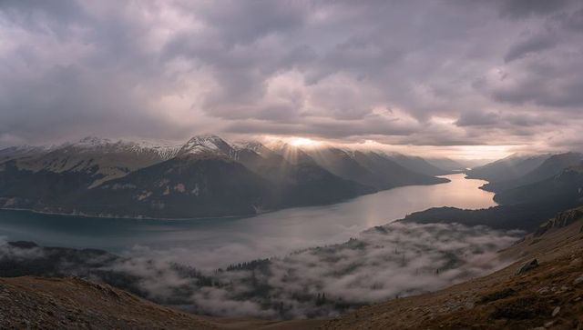 Sunbeams Breaking Through Clouds over Alpine Valley with Winding Lake and Mist at Sunrise