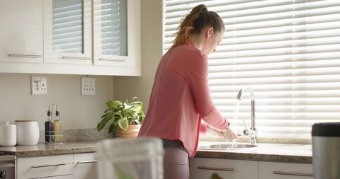 Young woman washing hands at sunlit kitchen sink with blinds and indoor plant