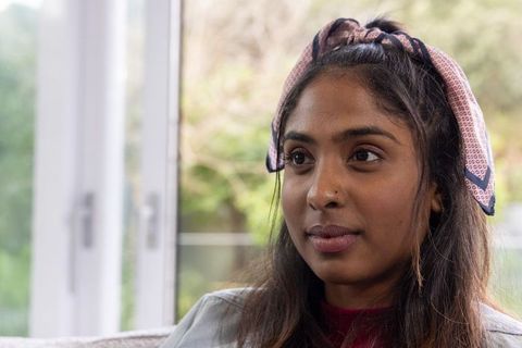 Indian woman sitting on sofa by window wearing patterned pink headscarf in natural light