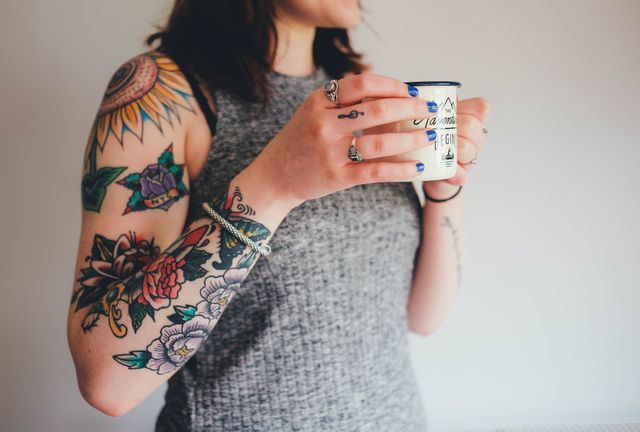 Young woman with colorful tattoos enjoying coffee while wearing grey tank top. Depicts casual, relaxed lifestyle and modern fashion. Useful for advertising tattoo parlors, coffee shops, and lifestyle brands targeting young audiences.