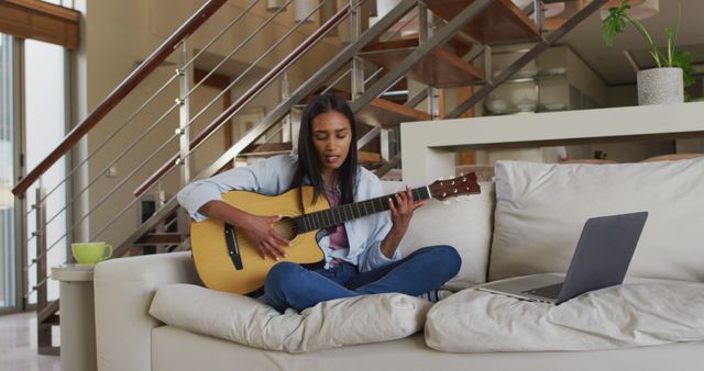 Young woman sits on a modern living room couch, practicing guitar while watching an online tutorial on a laptop. Wooden staircase and indoor greenery add a cozy feel. Ideal for use in articles about learning new skills from home, remote education, or lifestyle blogs emphasizing indoor activities.