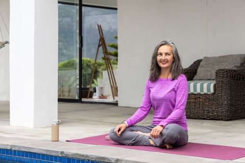 Middle-aged woman practicing yoga by pool on covered patio wearing purple top and gray pants