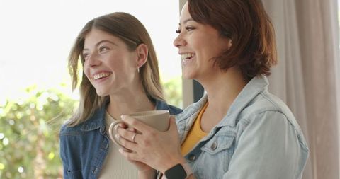 Sunlit friends sharing coffee by window, cozy denim morning, smiling and laughing together
