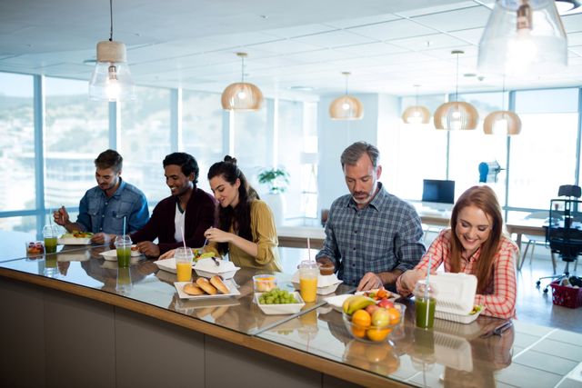 Diverse group of colleagues enjoying lunch together in a modern office setting. Ideal for use in articles or advertisements related to workplace culture, team building, corporate wellness programs, and healthy eating in the office.